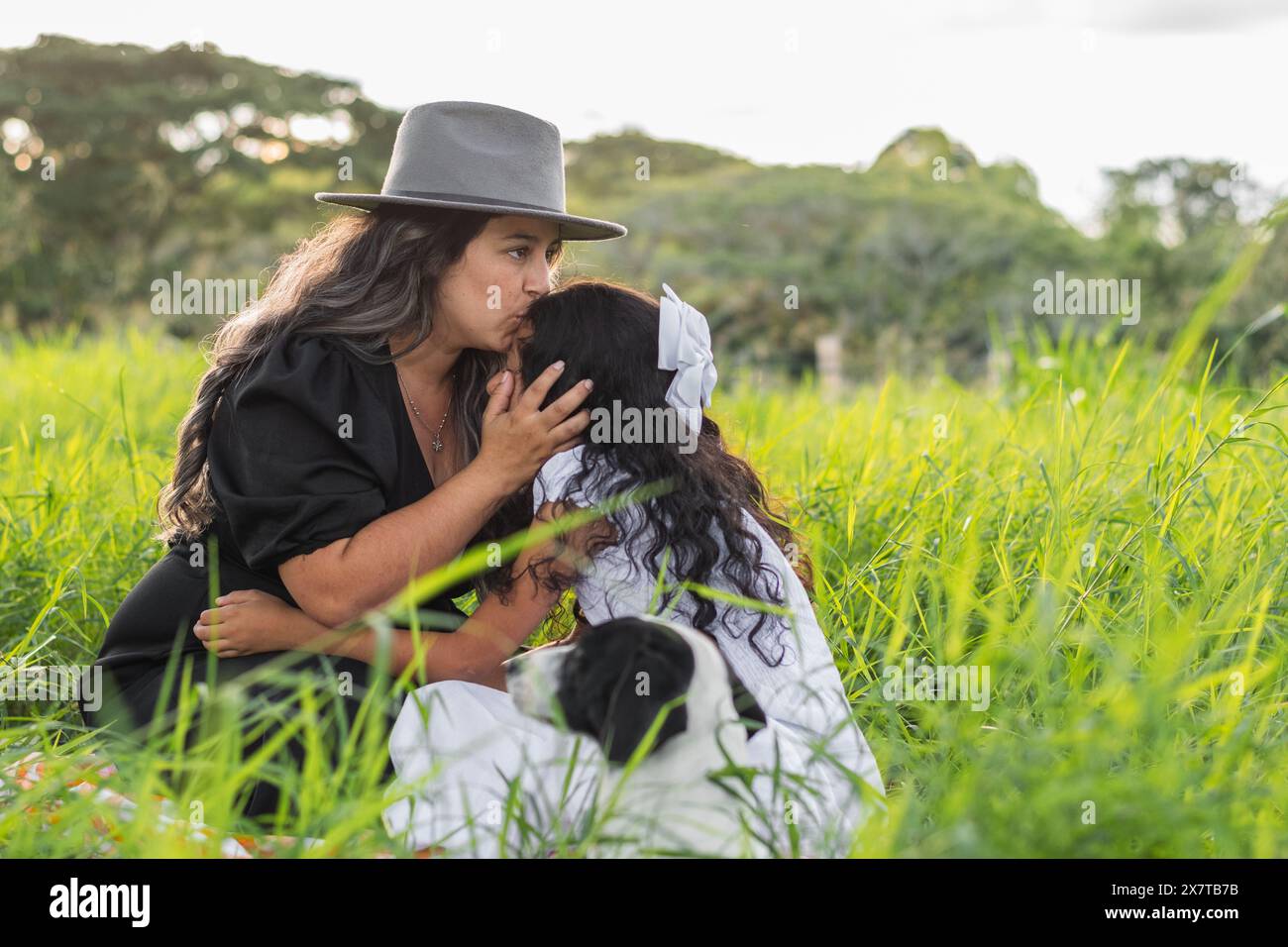 young latin mother kissing her little daughter's forehead while sitting on the grass Stock Photo