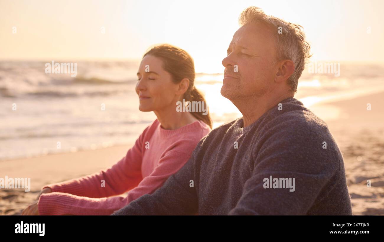 Peaceful Senior Couple Relaxing Sitting On Beach Shoreline With Closed Eyes At Sunrise Together Stock Photo
