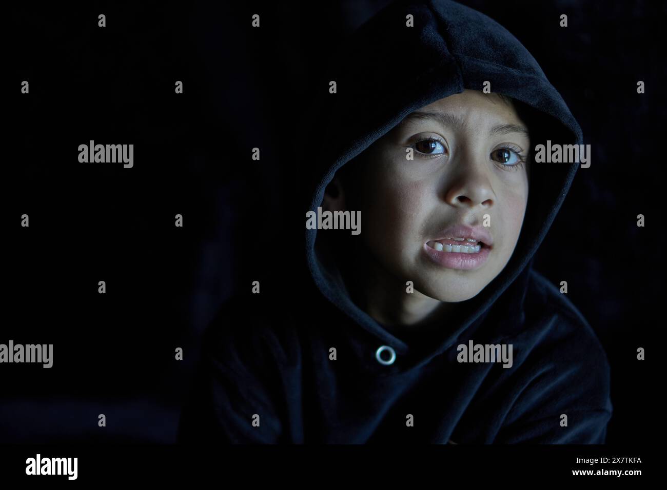 latino boy with black hood on his head and a confused expression on his face, black background Stock Photo