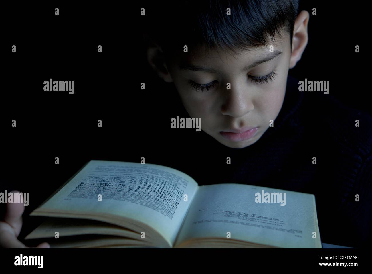 close-up portrait of intelligent Latino boy's face reading a book in deep concentration in the dark. Concept of gifted child Stock Photo