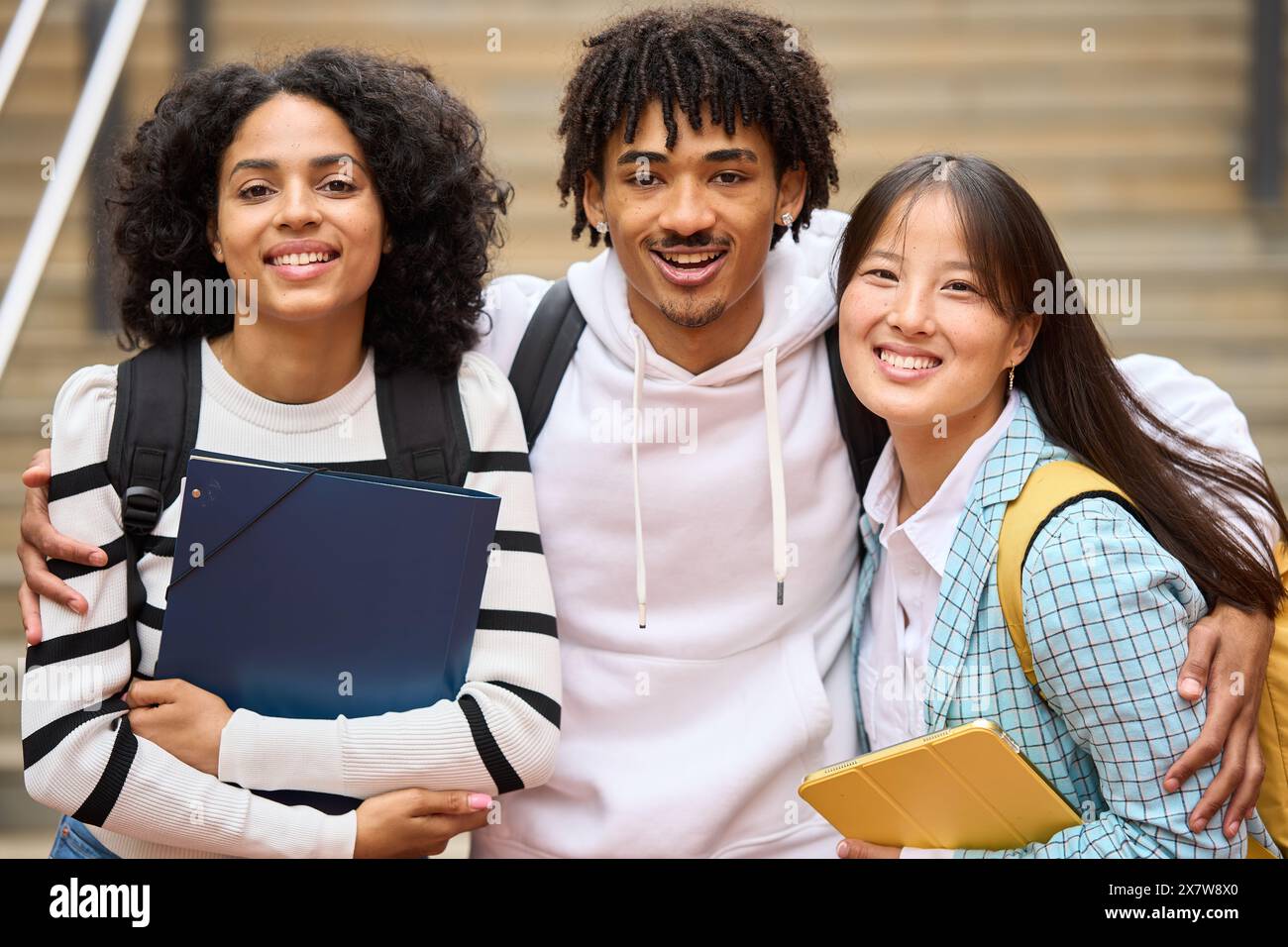 Three young students are smiling and hugging each other while holding backpacks, note pads and looking at camera Stock Photo