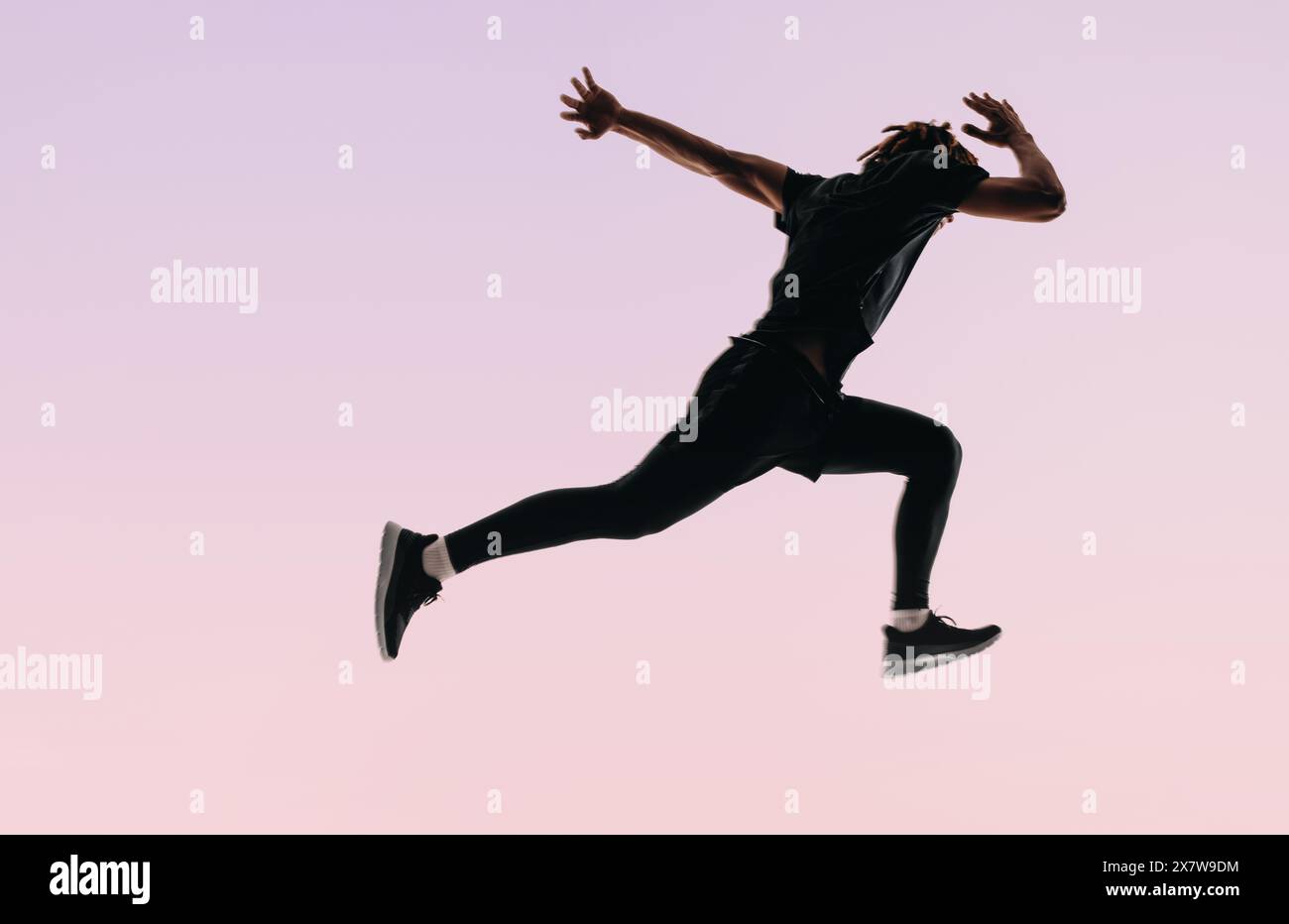 Fit man with dreadlocks jumps mid-air, showcasing his dedication to fitness. Against a pink background, he practices running and sprints in sportswear Stock Photo