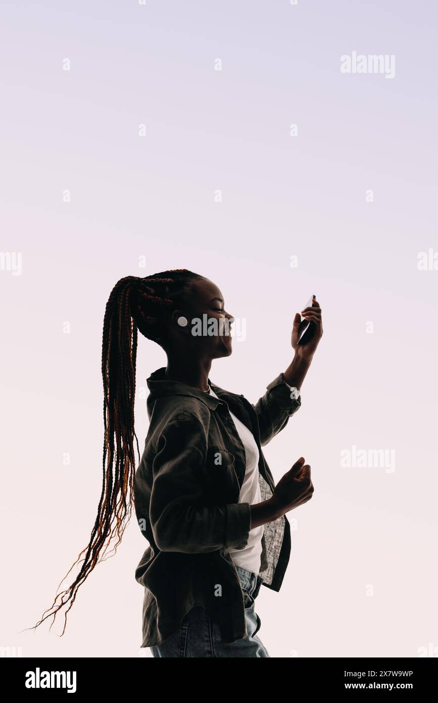 Young woman with braided hair dances happily in a studio, holding a smartphone and wearing wireless earbuds. She expresses herself through movement. Stock Photo