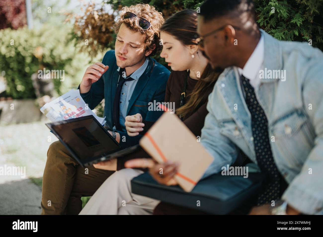 Three diverse professionals collaborating outdoors with a laptop and documents Stock Photo