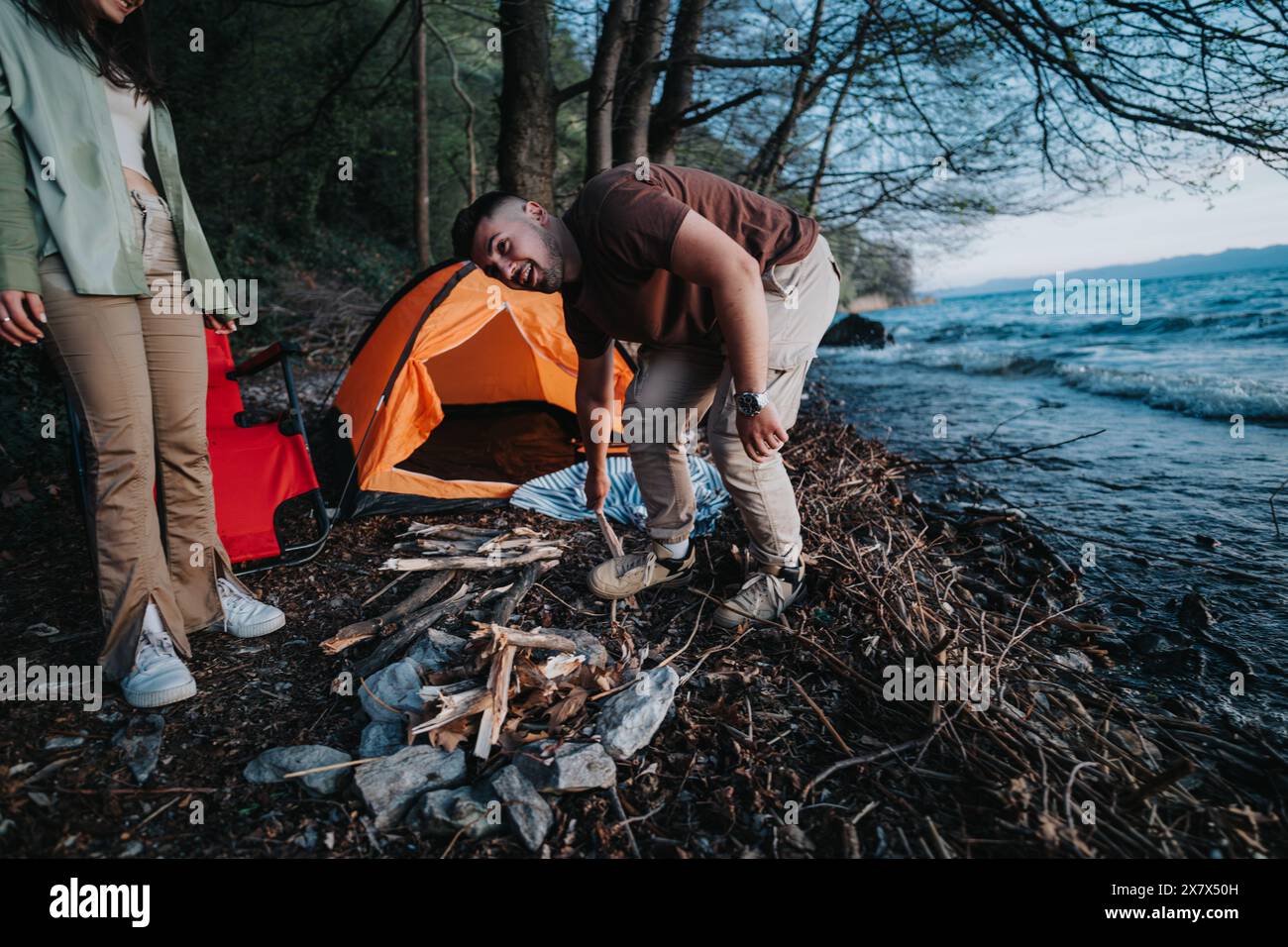 Friends Setting Up Campsite By A Lake With A Tent And A Fire Pit Stock Friends Setting Up Campsite By A Lake With A Tent And A Fire Pit Stock