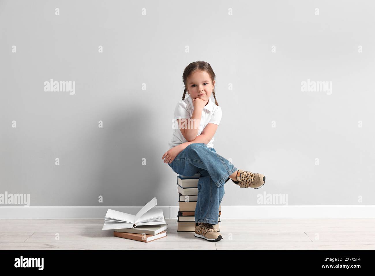 Cute little girl sitting on stack of books near light grey wall Stock ...