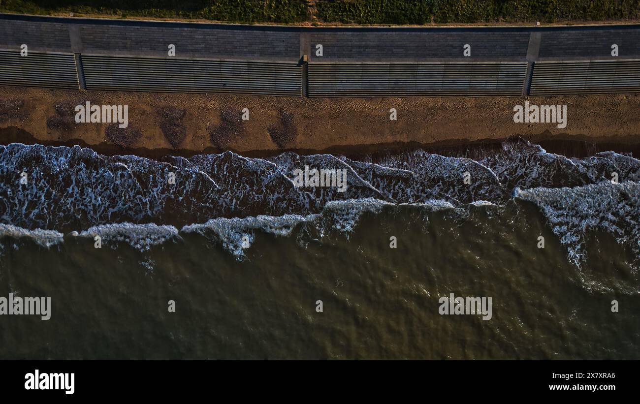 Aerial view of ocean waves crashing onto a sandy beach with a concrete seawall in the background. Stock Photo