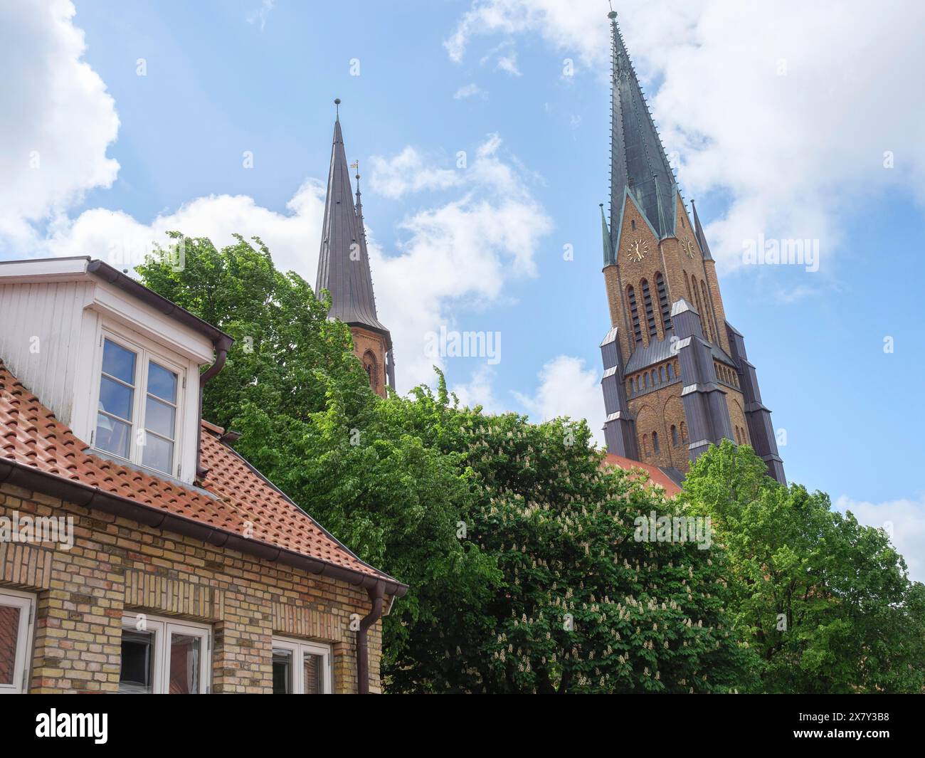 A tall church tower dominates the scenery next to a brick building, surrounded by trees and clouds, large church towers with trees and historic houses Stock Photo