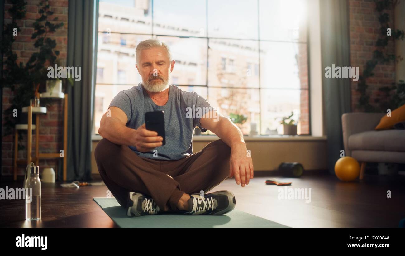Portrait of a Strong Athletic Fit Senior Man Using Smartphone in Between Workouts During Morning Exercises at Home in Sunny Apartment. Healthy Lifestyle, Fitness, Recreation, Wellbeing and Retirement. Stock Photo