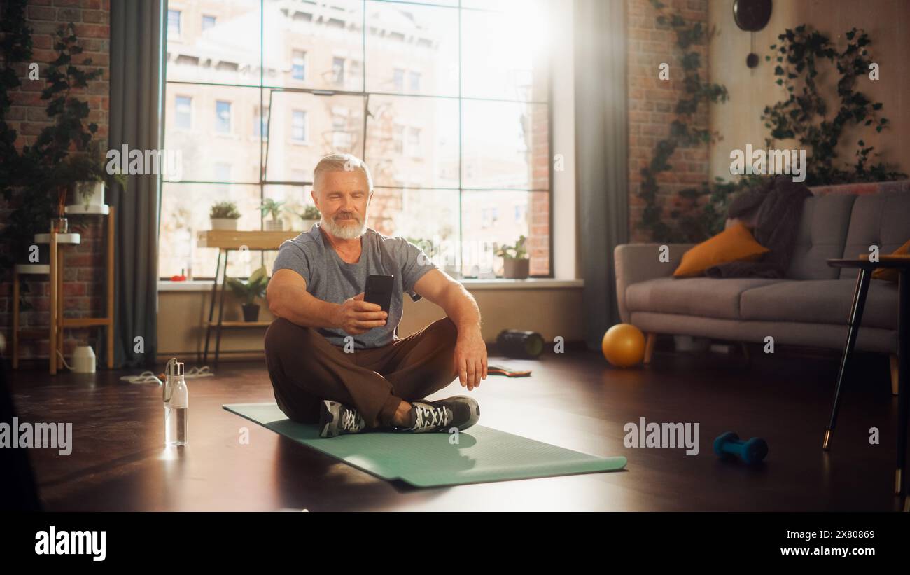 Strong Athletic Fit Middle Aged Man Using Smartphone in Between Workouts During Morning Exercises at Home in Sunny Apartment. Healthy Lifestyle, Fitness, Recreation, Wellbeing and Retirement. Stock Photo