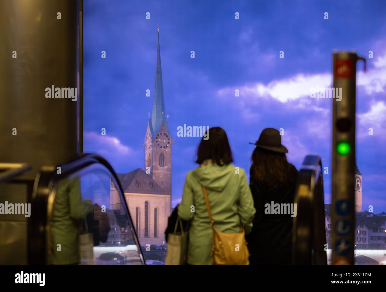 Zurich, Switzerland - May 16, 2024: Two individuals on escalator, against urban view of Zurich with historic church and dramatic sky. Stock Photo