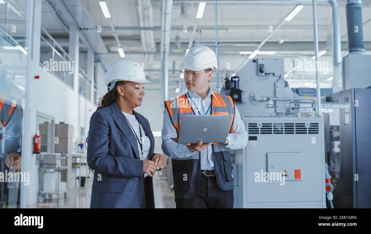 Two Professional Heavy Industry Engineers Walking at a Factory in Safety Uniform and Hard Hats, Discussing Work on Laptop Computer. Asian Specialist and African American Technician at Work. Stock Photo