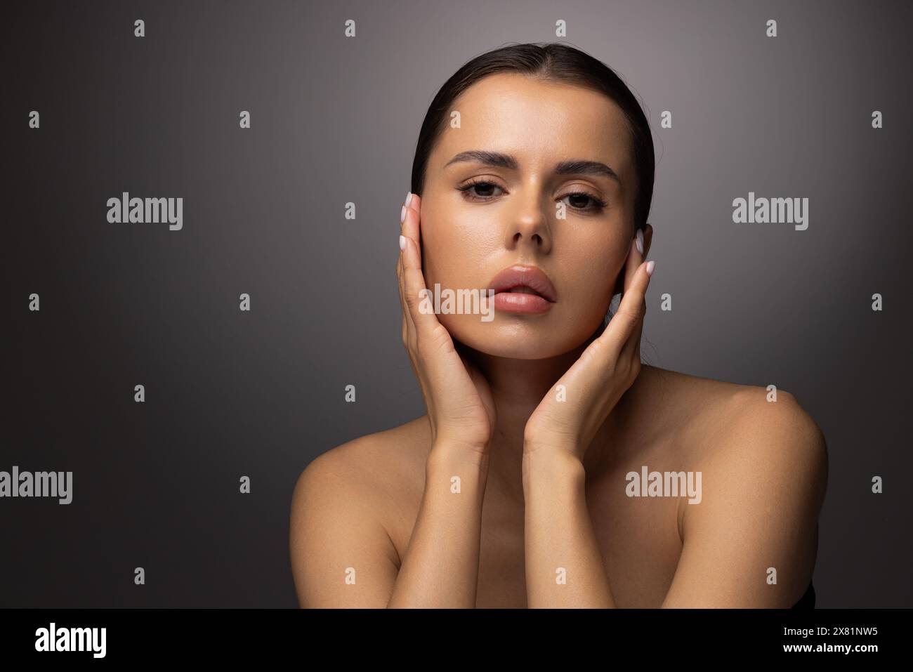 Beauty concept, head and shoulders portrait of beautiful model with nude make-up looking at camera, touching face, closeup. Brunette woman in studio w Stock Photo