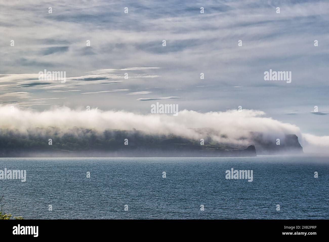 A serene coastal landscape with a calm sea and a distant landmass partially covered by low-lying clouds. The sky is overcast with a mix of clouds and Stock Photo