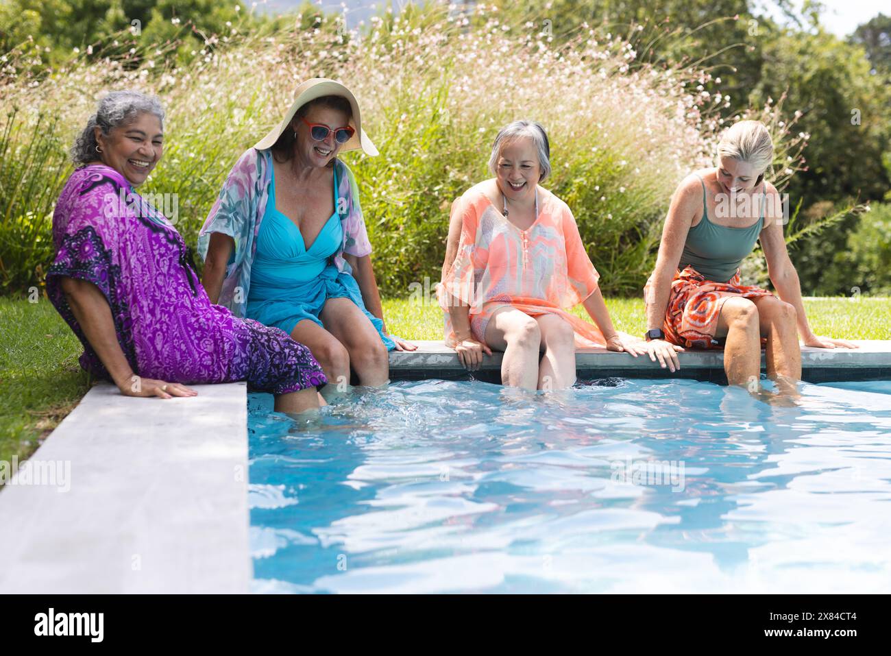Outdoors, diverse senior female friends sitting by pool, dipping feet in water, copy space Stock Photo