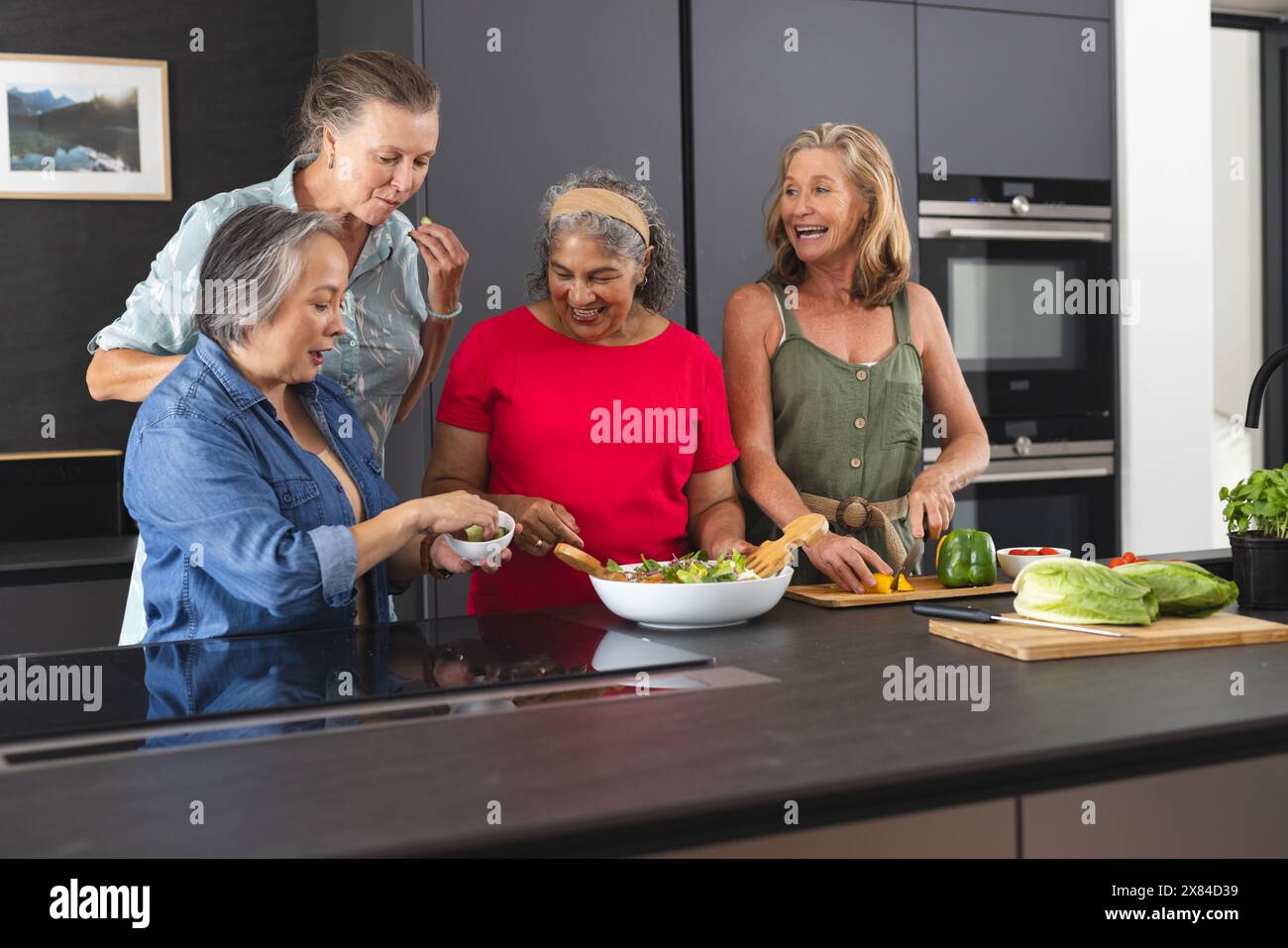 Diverse senior female friends preparing salad at home, laughing together Stock Photo