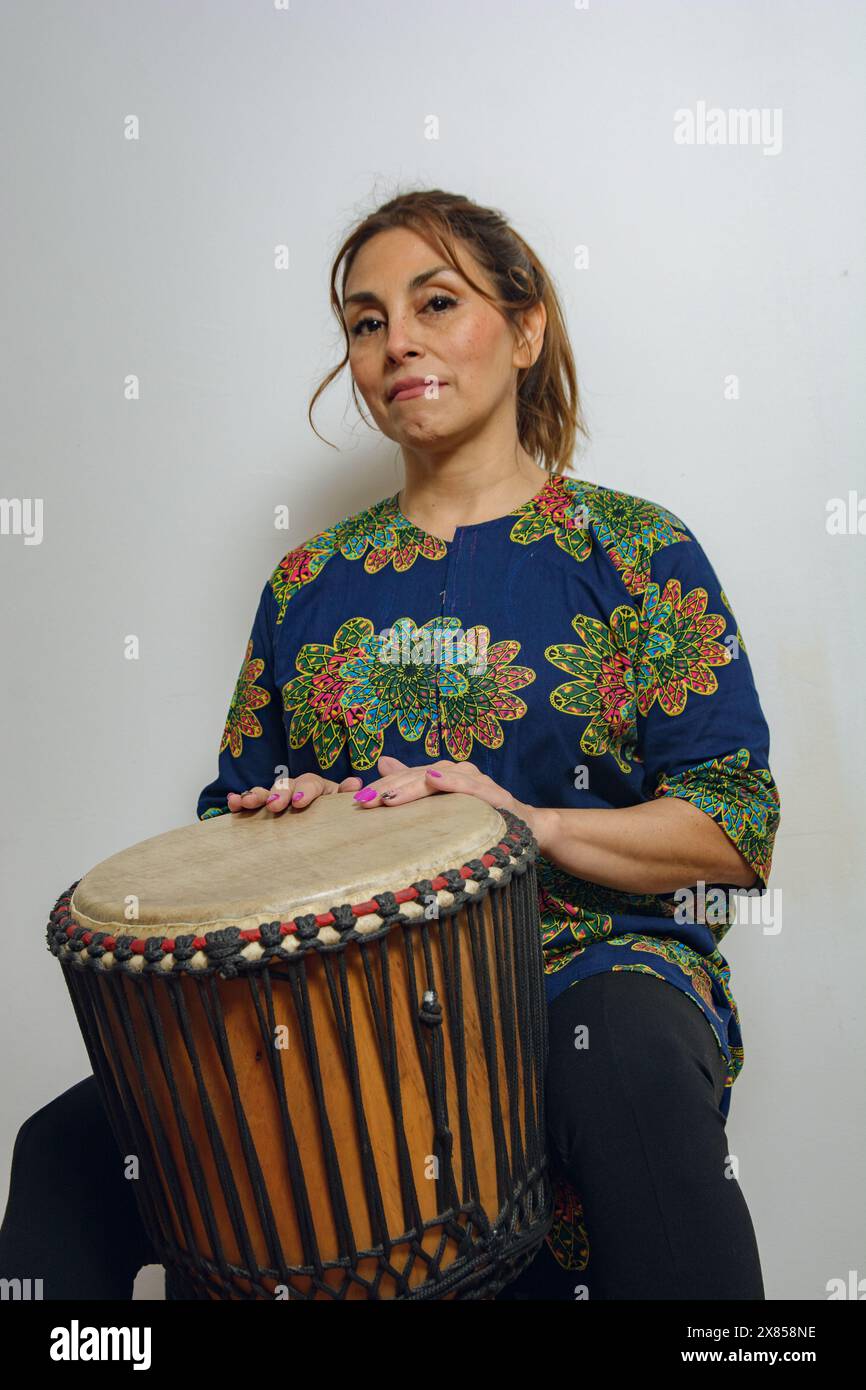 An Adult Latin Woman wearing an African style clothing is sitting playing a yembe drum looking at camera. The drum is made of wood and has a black and Stock Photo