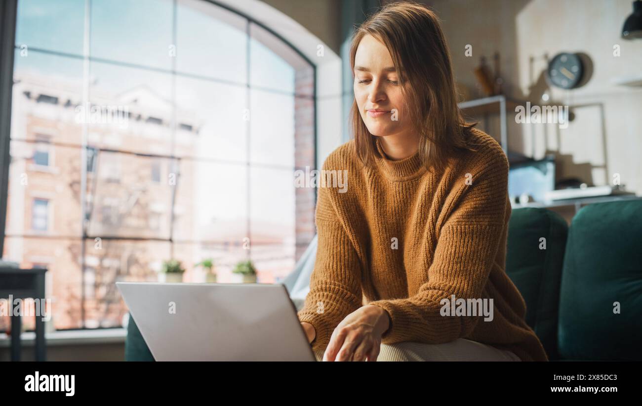 Smiling Young Woman Working from Home on Laptop Computer in Her Sunny Cozy Apartment, Relaxes and Leans Back on a Sofa. Successful Creative Female Entrepreneur Happy After Finishing Remote Work Stock Photo