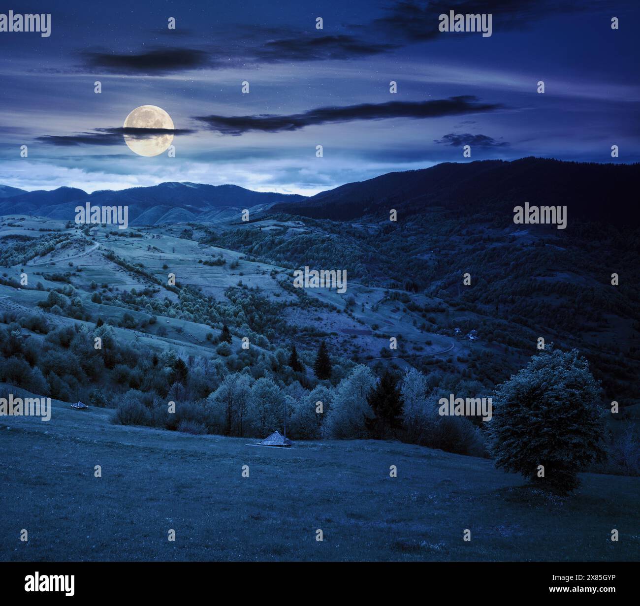 rural mountain landscape in spring at night. grass and trees on the hills rolling through the green valley in to the distant ridge beneath a cloudy sk Stock Photo