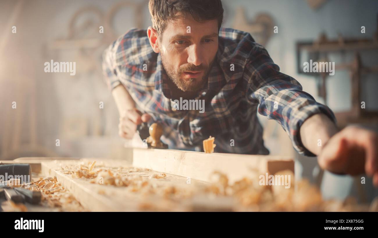 Handsome Young Artisan Craftsman in Checkered Shirt Using Hand Plane to Shape a Wood Bar. Carpenter Working on a Project in a Loft Studio with Tools on Walls. Stock Photo
