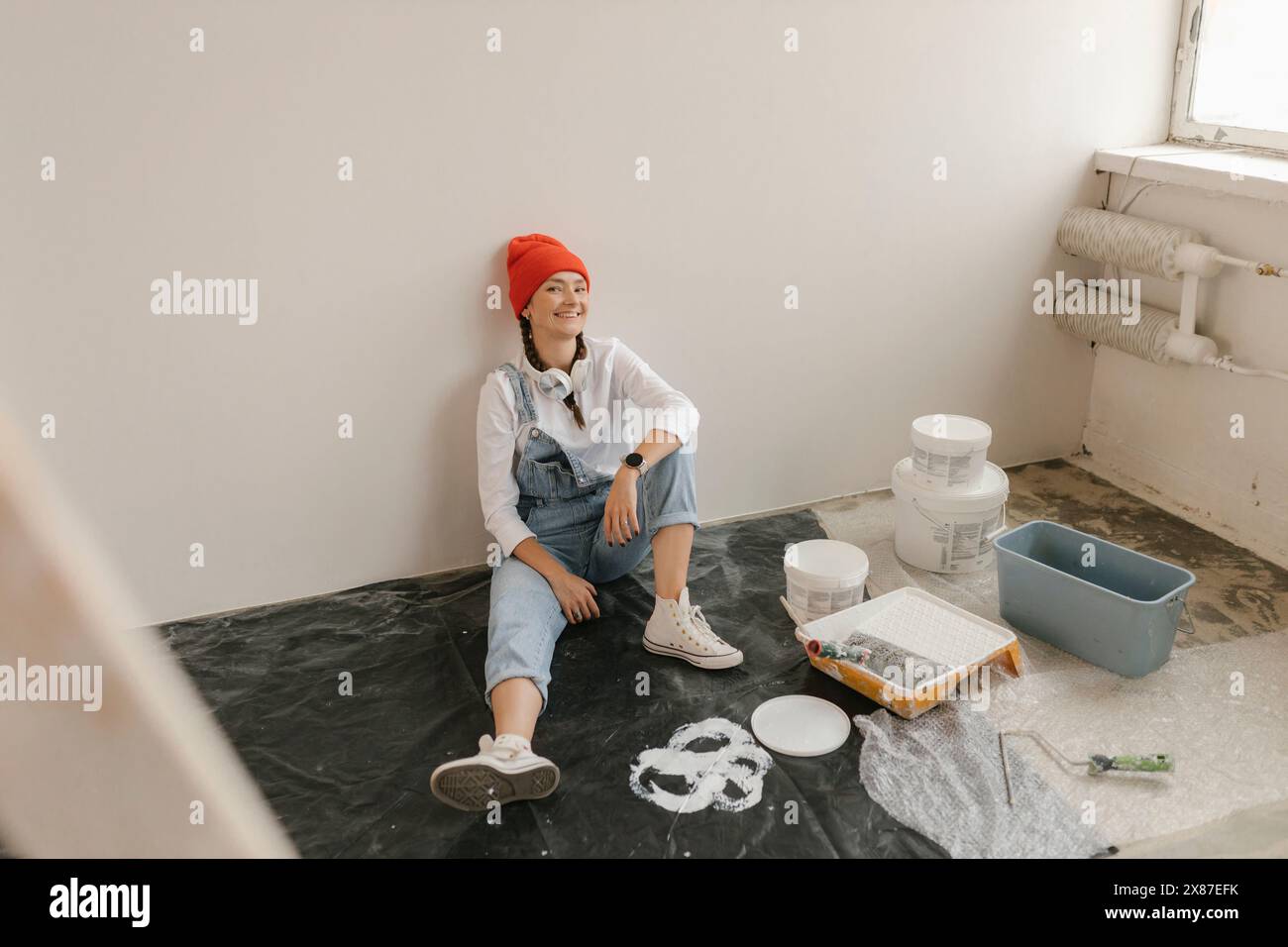 Smiling woman leaning on white wall sitting by paint equipment in new apartment Stock Photo