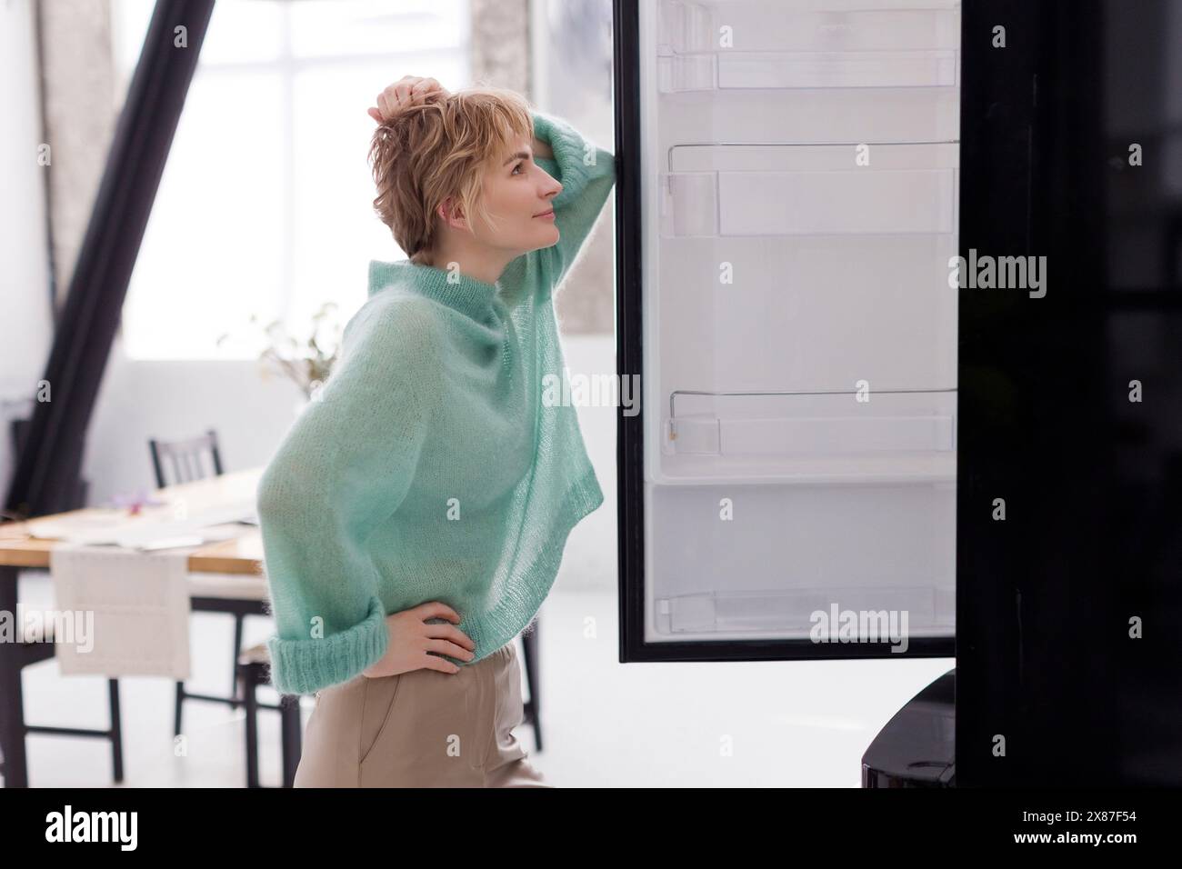 Blond woman with hand on hip leaning on refrigerator door at home Stock Photo