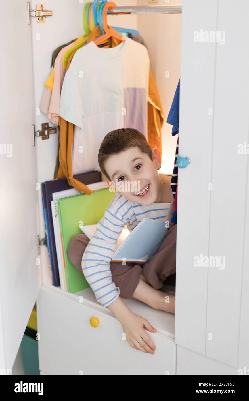 Smiling boy sitting cross-legged with book in wardrobe at home Stock Photo