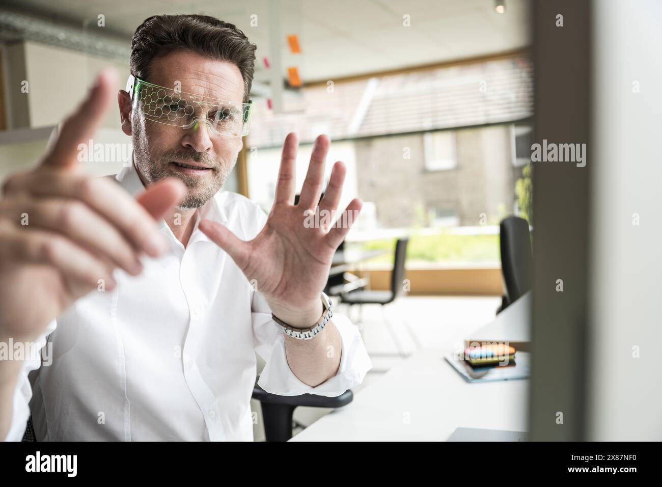 Confident businessman gesturing and wearing smart glasses sitting at office Stock Photo