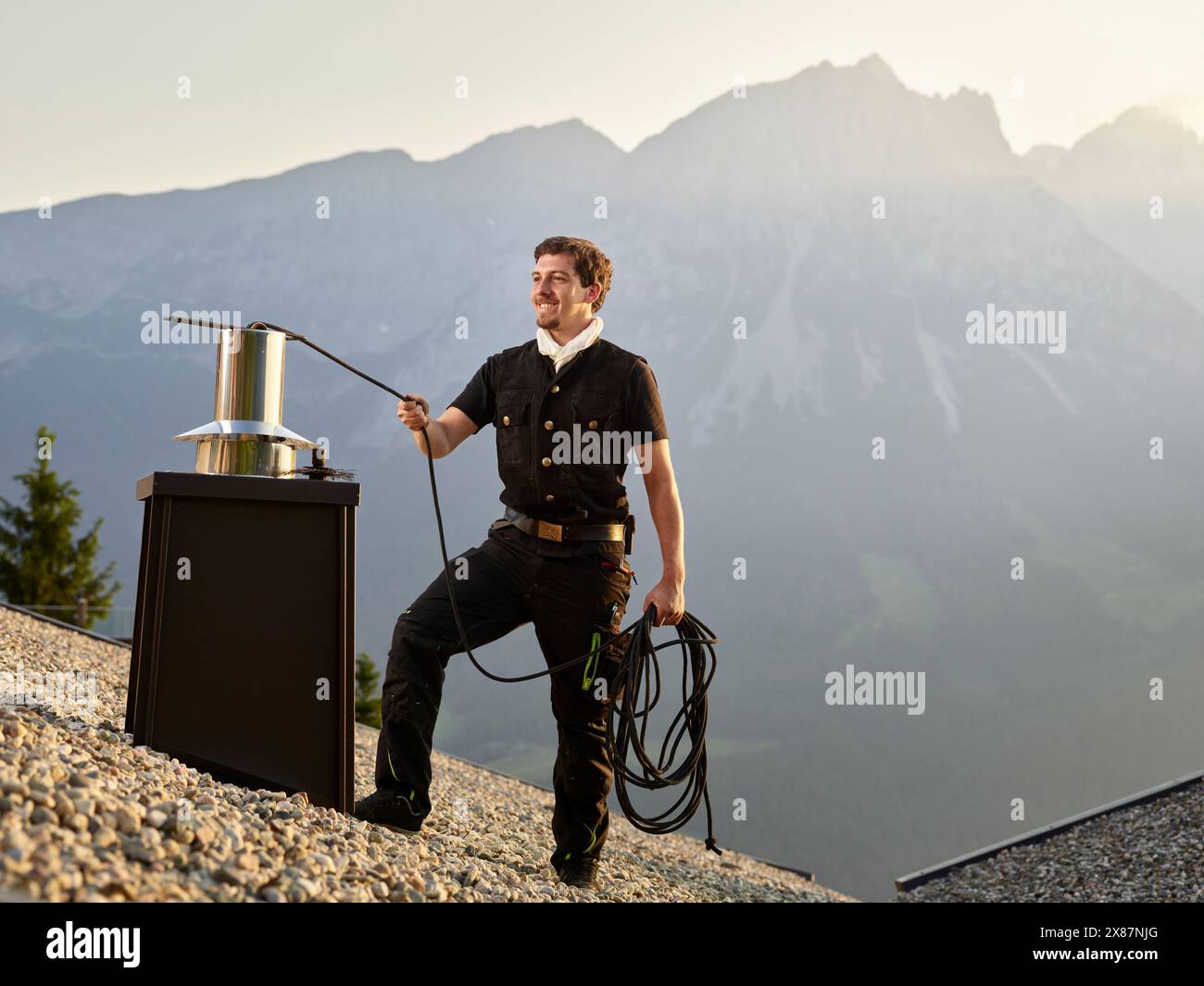Smiling chimney sweeper servicing chimney on rooftop at sunrise Stock Photo