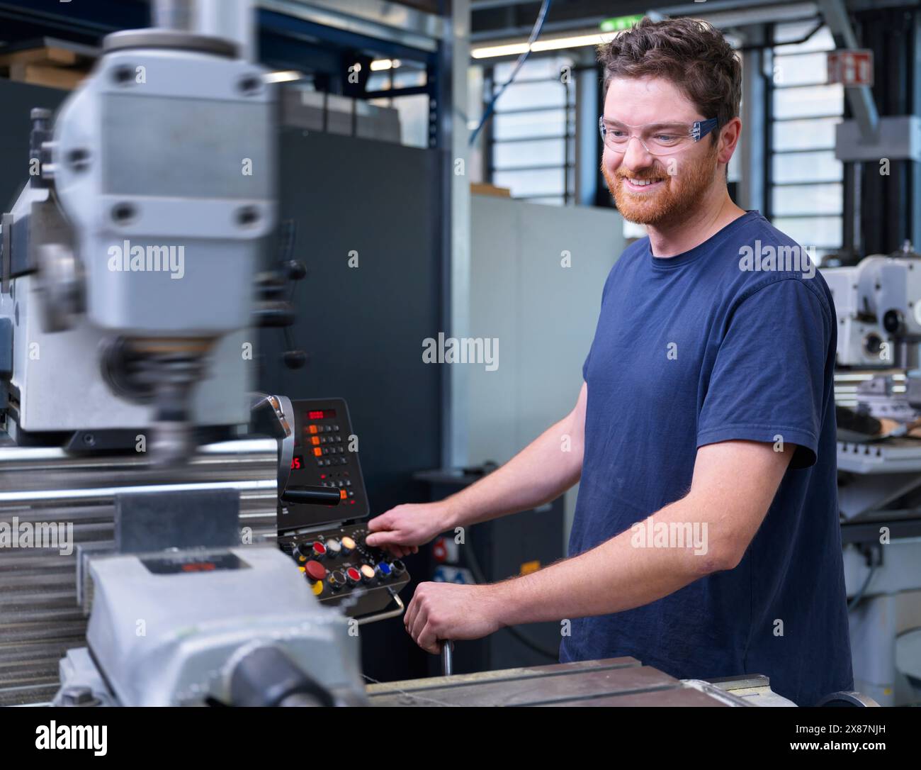 Smiling engineer in blue t-shirt operating CNC machine at factory Stock Photo