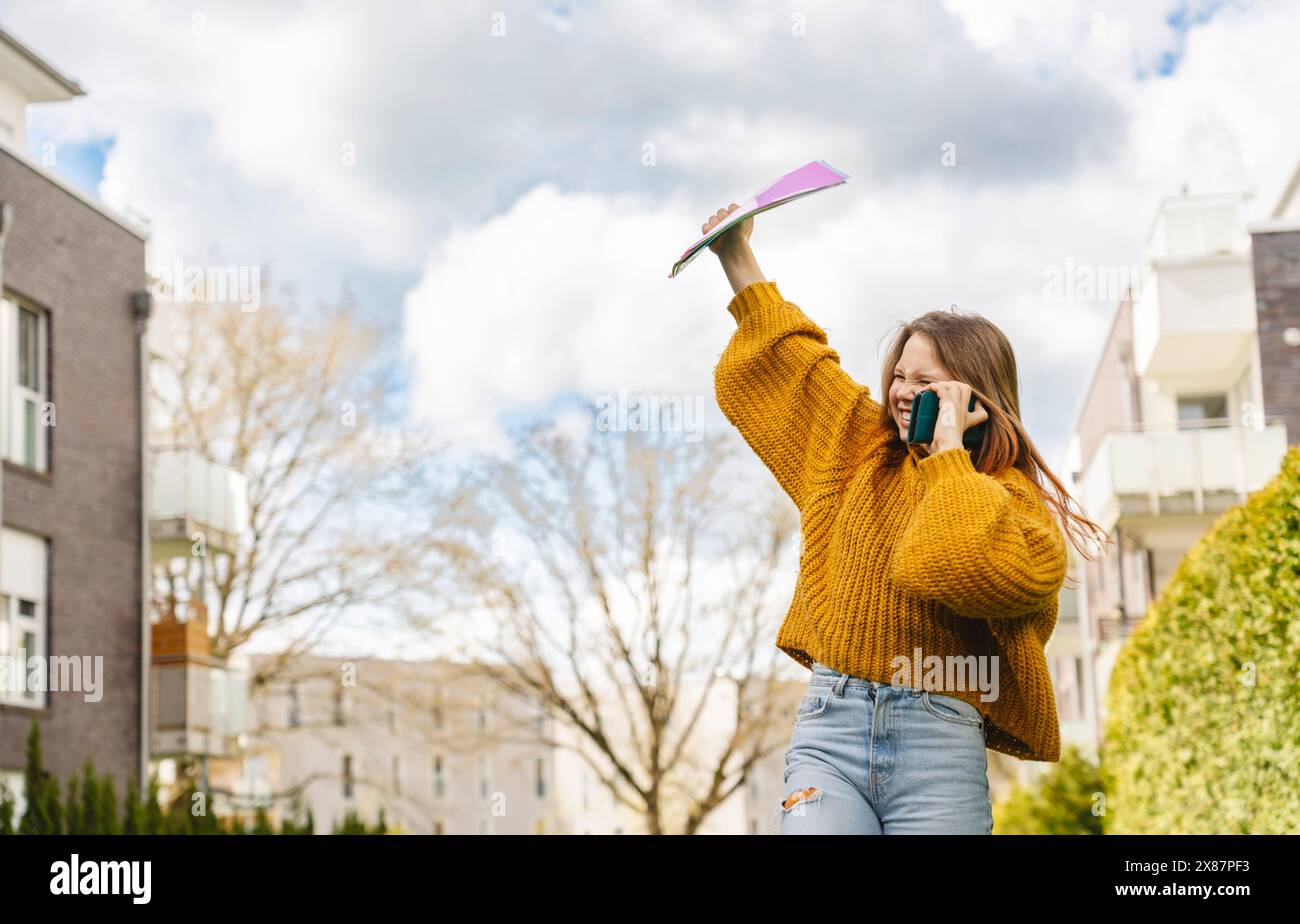 Cheerful girl talking on smart phone and holding folders in hand under cloudy sky Stock Photo
