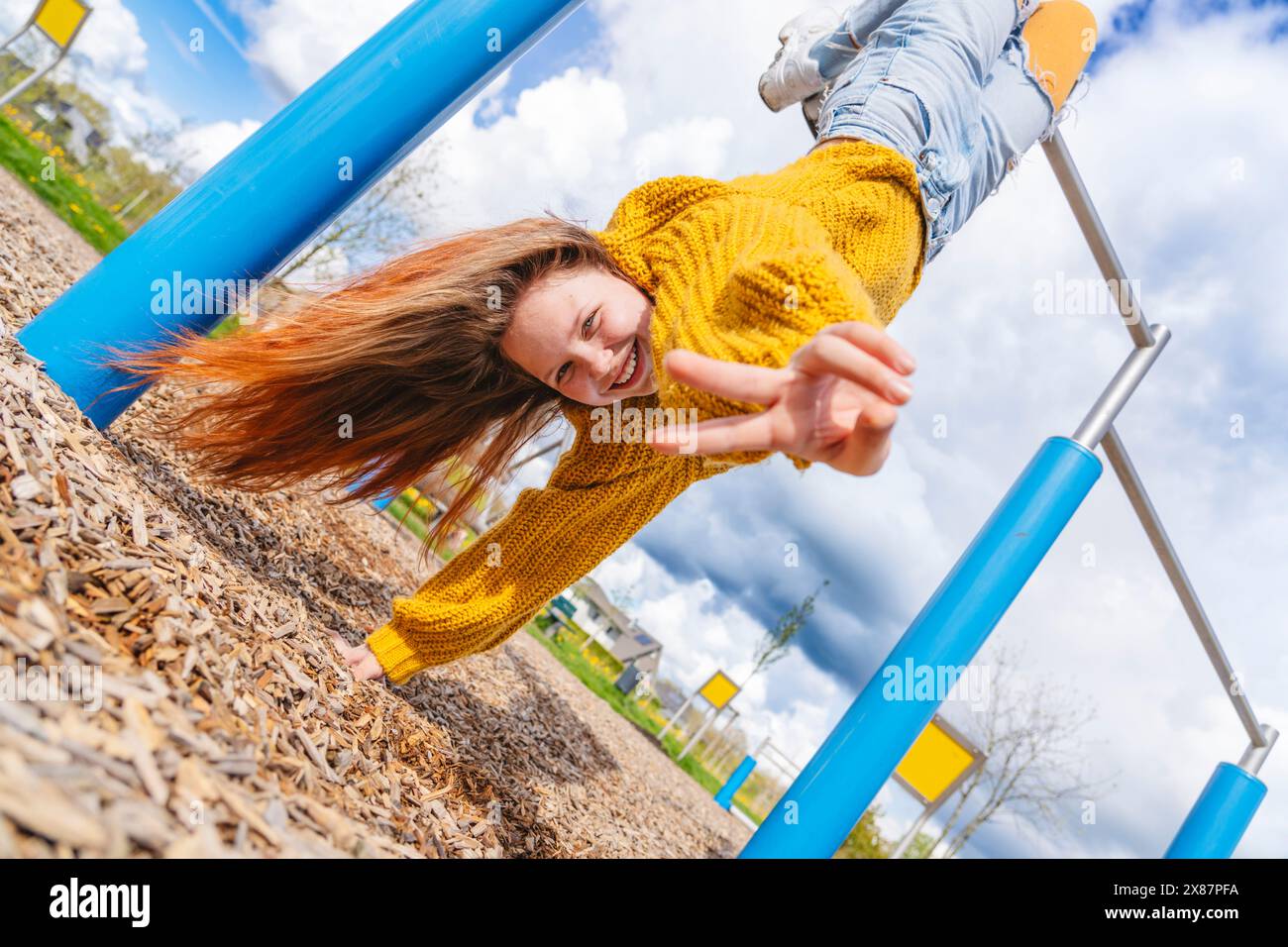 Cheerful girl hanging upside down on gymnastics bar and showing peace sign Stock Photo