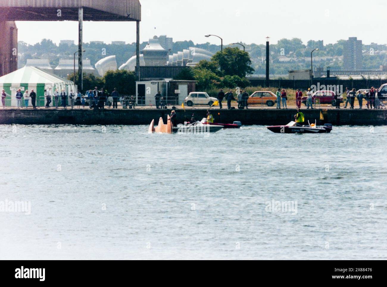 Powerboat racing event in Royal Victoria Dock, London, UK, in 1986, before the area was developed. Grand Prix of London. The Thames Barrier visible beyond. Crashed, partially submerged powerboat with rescue boats on scene and old cars of the 1980s era Stock Photo