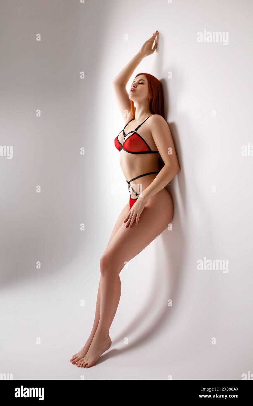 Young sexy redhead woman in see-through red lingerie leaning on a white  wall in studio and attractively posing with her hand at her hip and  touching t Stock Photo - Alamy