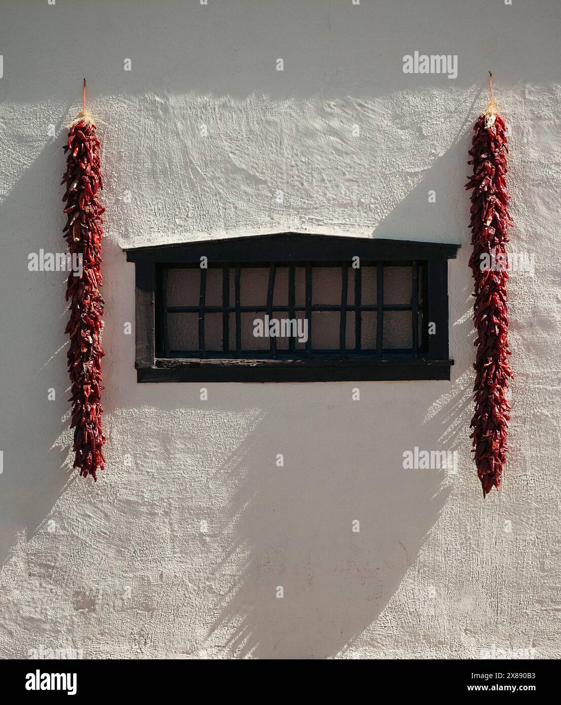 Side lighting of red chili ristras hanging on white wall with black wooden window on exterior of traditional Southern New Mexican abode in Mesilla, NM Stock Photo