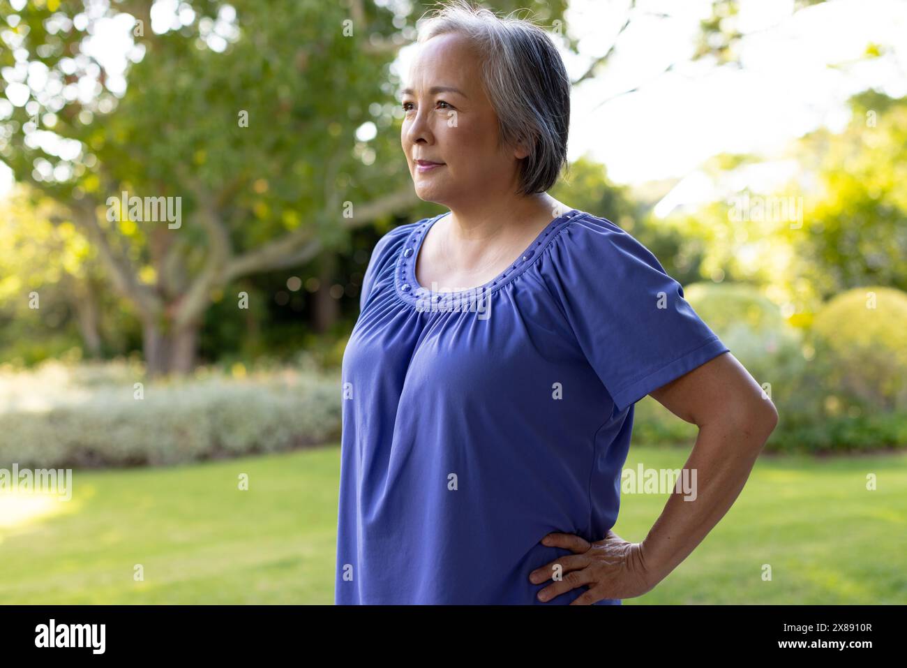 Asian middle-aged woman stands in garden, hands on hips, gazes sideways Stock Photo