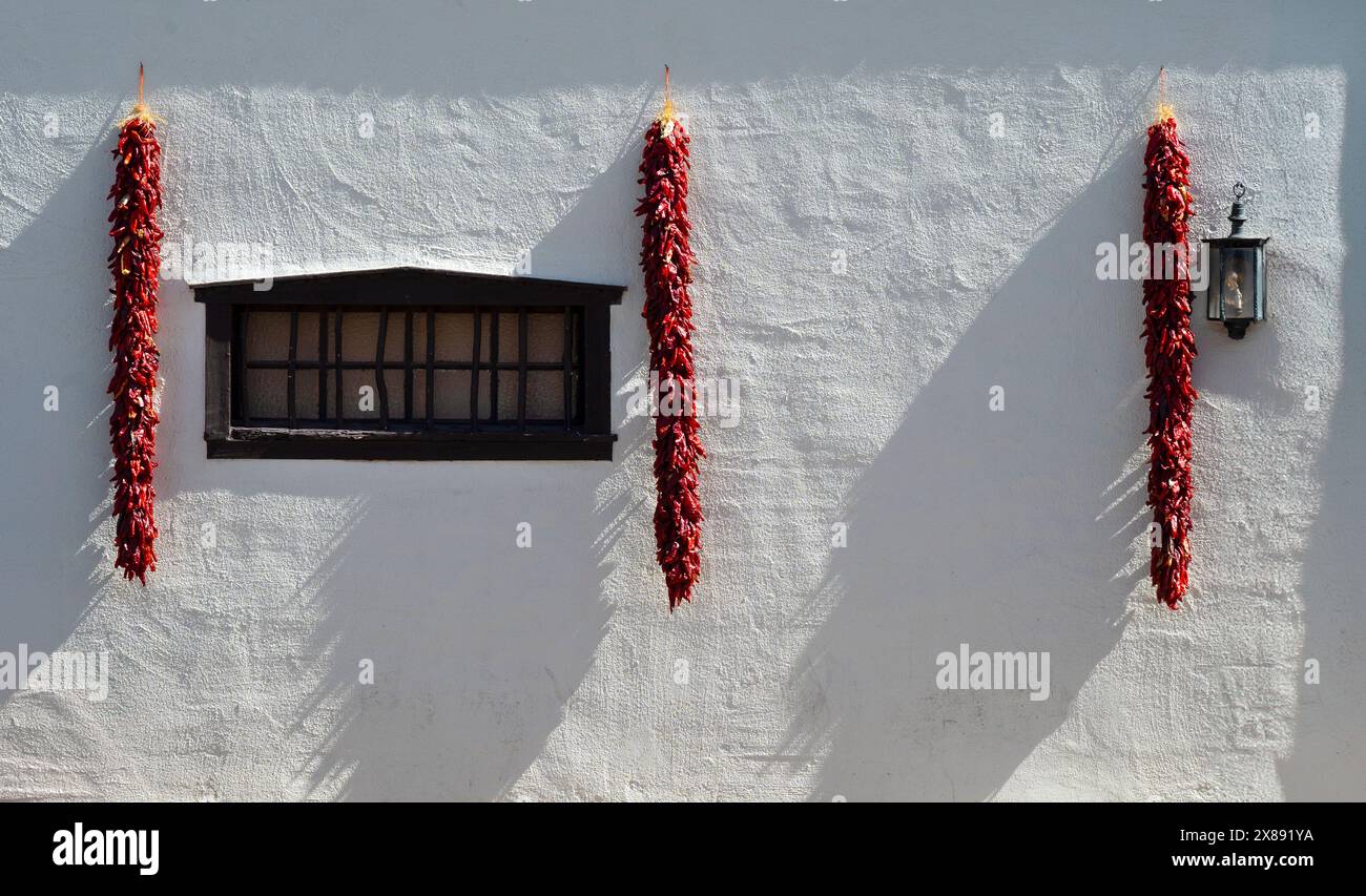 Side lighting of red chili ristras hanging on white wall with black wooden window on exterior of traditional Southern New Mexican abode in Mesilla, NM Stock Photo