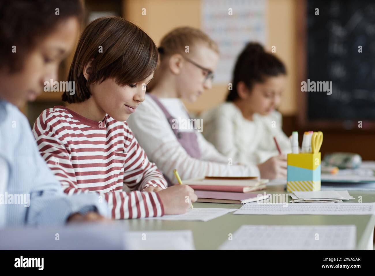 Selective focus shot of Caucasian boy wearing casual clothes sitting at table with his classmates doing writing exercise Stock Photo