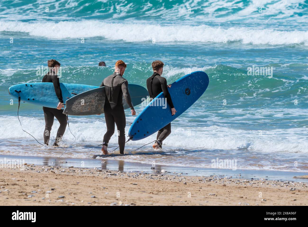 Three friends carrying their surfboards walking into the sea on Fistral Beach in Newquay in Cornwall in the UK. Stock Photo