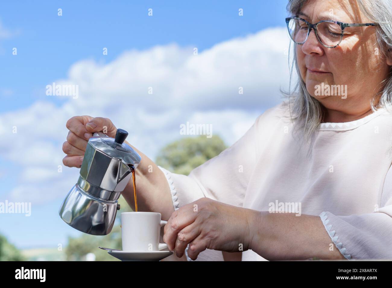 close-up of a woman serving herself coffee at a table in a field with a blue sky with clouds in the background Stock Photo