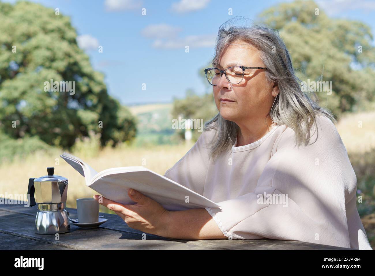 a woman reads a book at a table in a field while drinking a cup of coffee in the background trees and a blue sky with clouds. Stock Photo