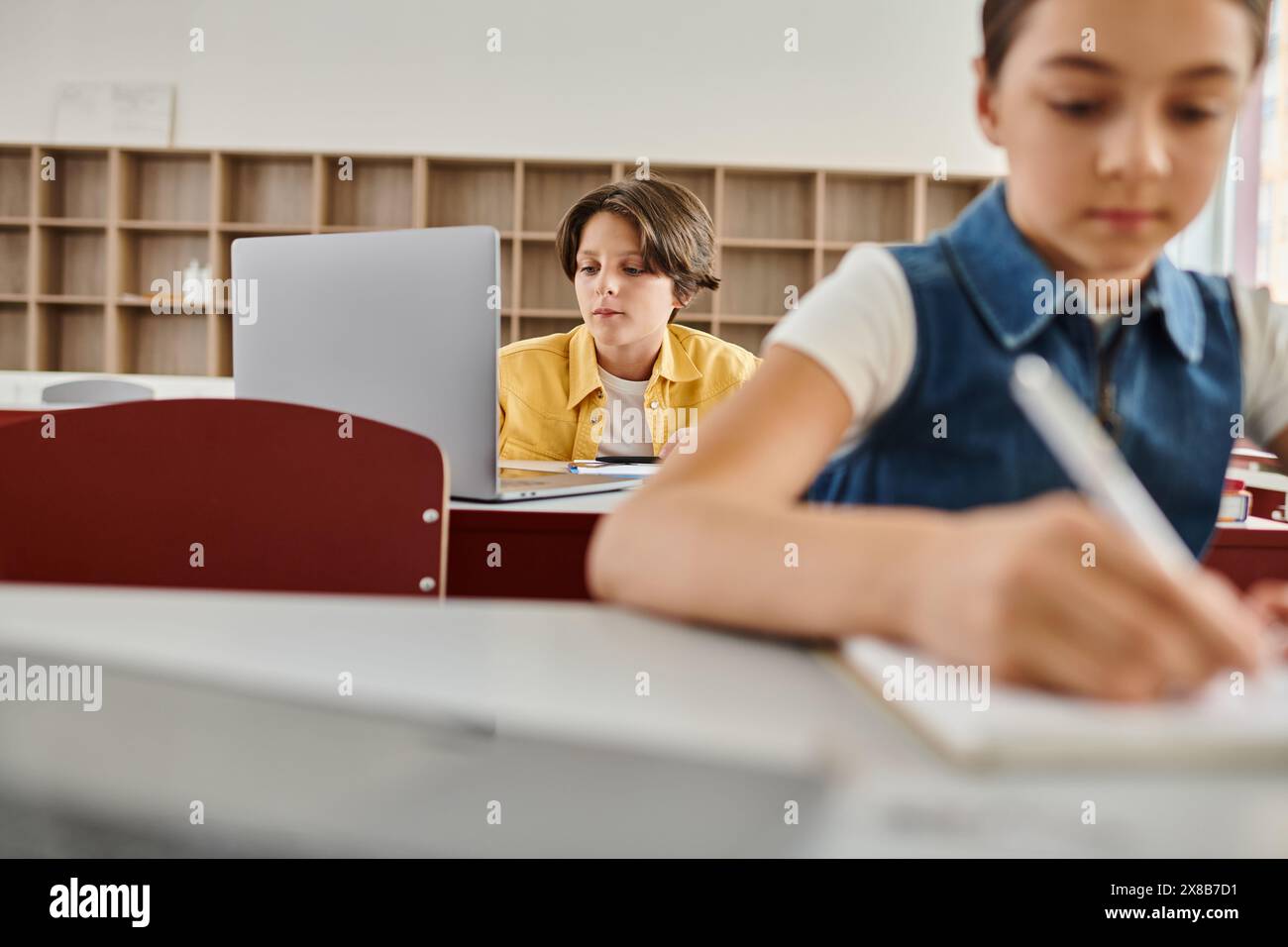 A young boy sits at a desk in a bright classroom, focused on laptop ...