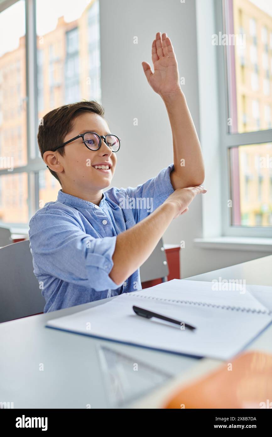 A young boy sits at a desk in a bright, lively classroom, raising his ...