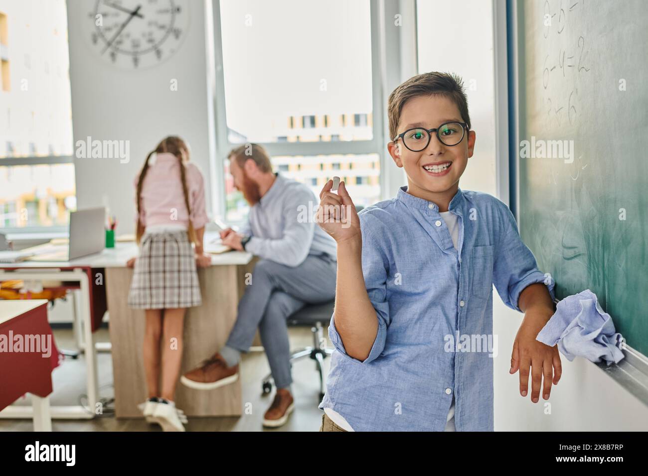 A young boy stands attentively before a blackboard in a busy class ...