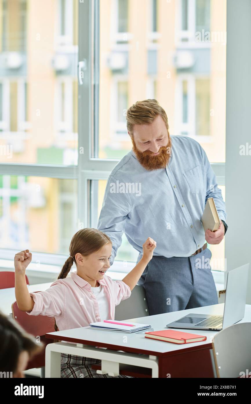 A man stands beside a little girl in front of a laptop computer, engaging in a lively tech-savvy learning session in a colorful classroom. Stock Photo