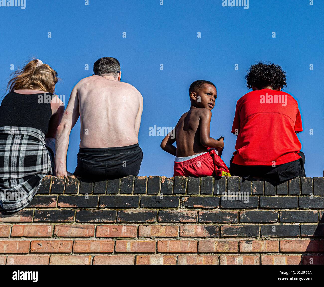 men and women and sitting on a wall facing away and a black child  without shirt looking behind . Stock Photo