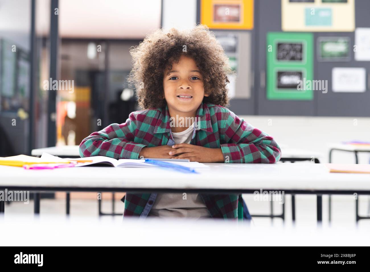 In school, young biracial boy leaning on a desk in a classroom, smiling ...