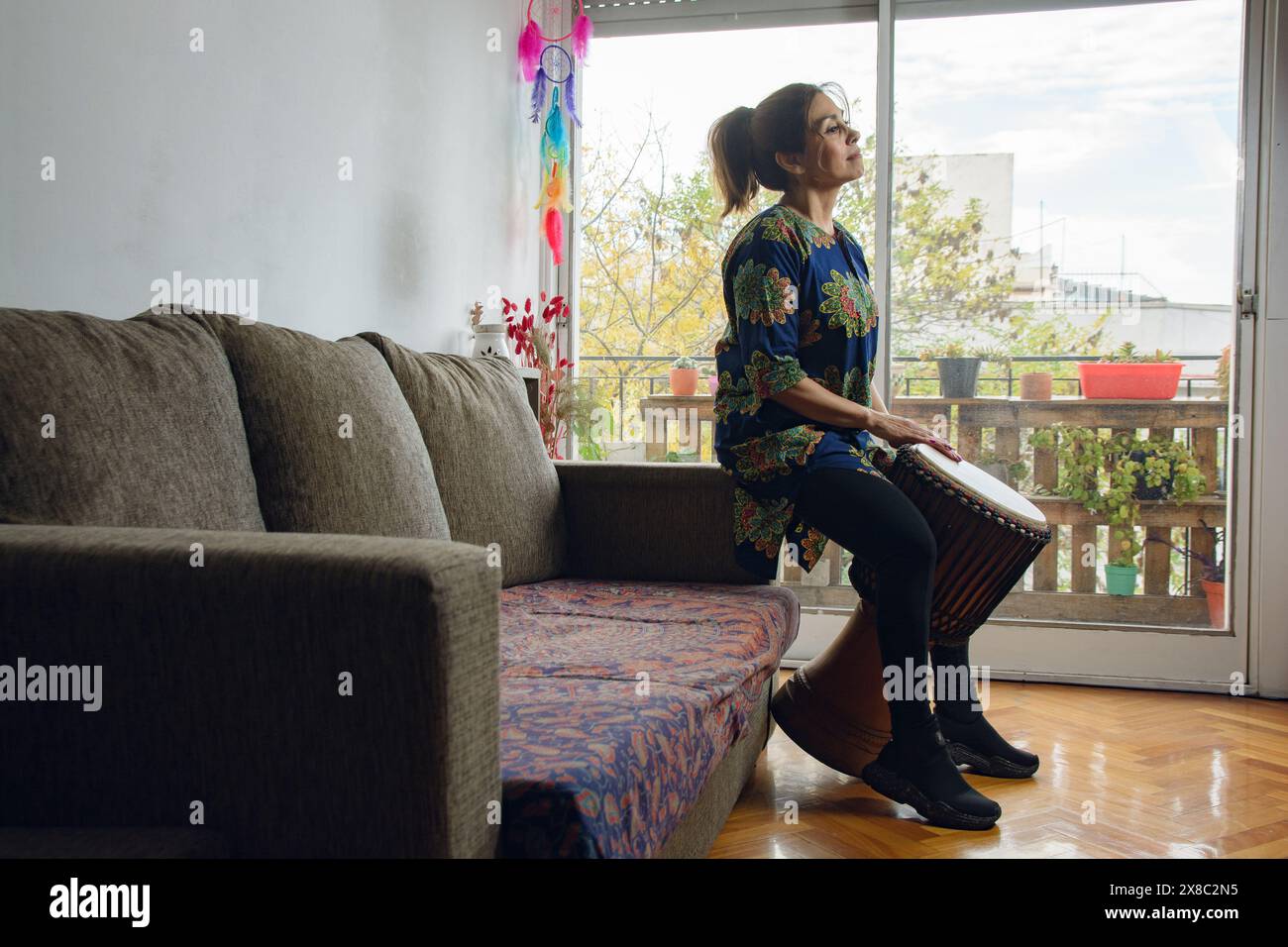 An Adult Latin Woman at home sitting on the couch playing the djembe drum, making music and looking up looking for inspiration. the balcony is in the Stock Photo