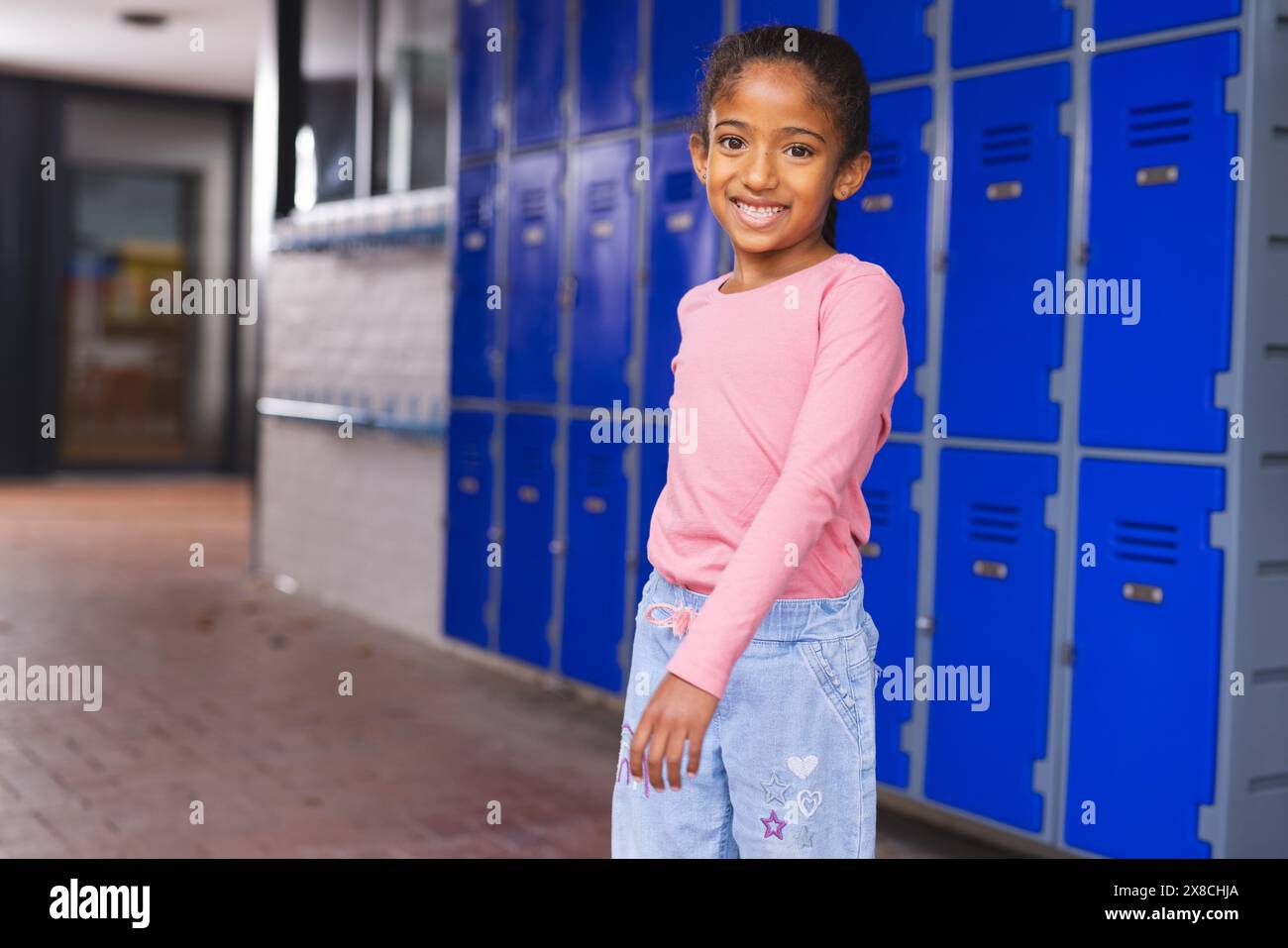 In school, young biracial girl is standing by blue lockers with copy space Stock Photo