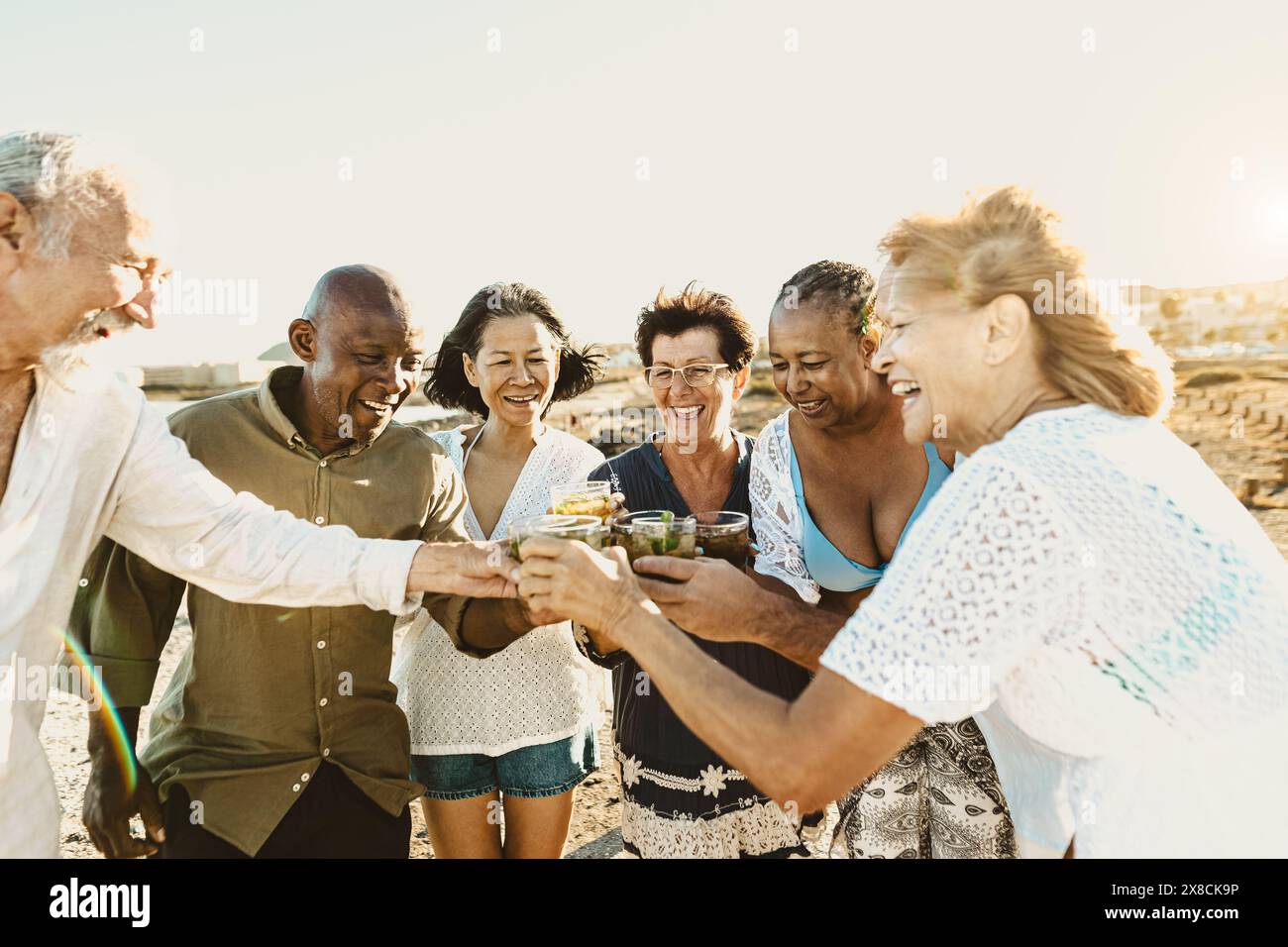 Happy multiracial senior friends having fun drinking and toasting mojitos on the beach during sunset time - Elderly people enjoying summer holidays Stock Photo